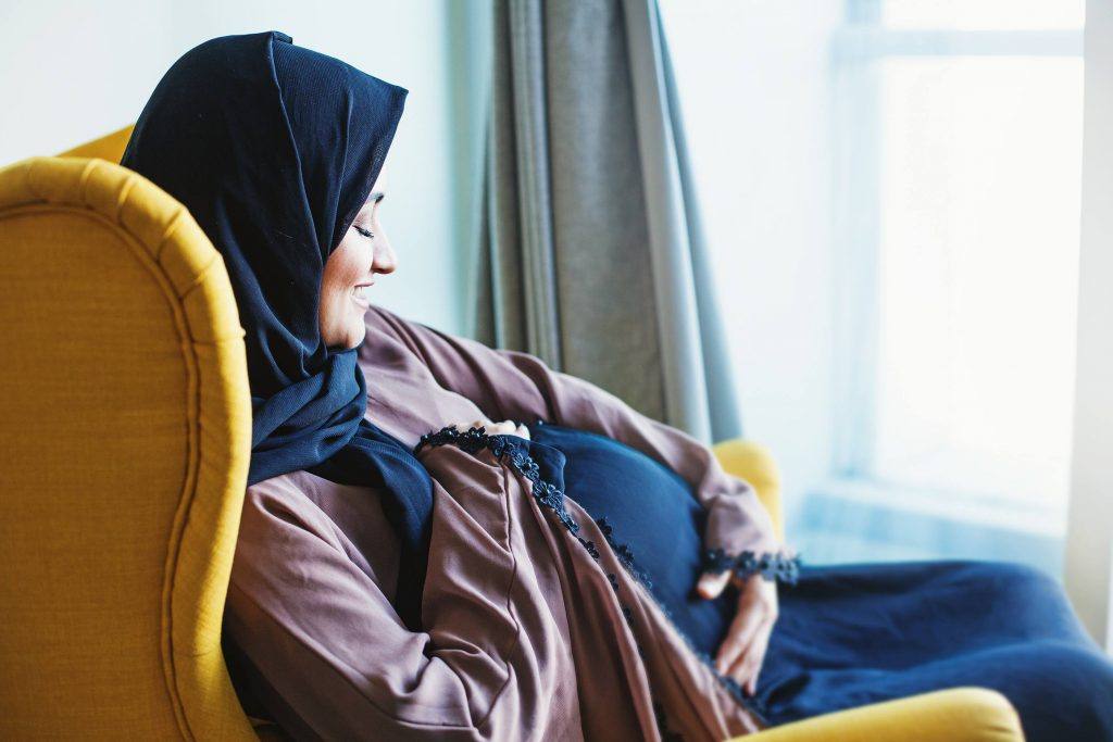 امرأة حامل شابة مبتسمة ترتدي الحجاب الشرعي، وتنظر إلى بطنها الحامل وتتلمَّسها.