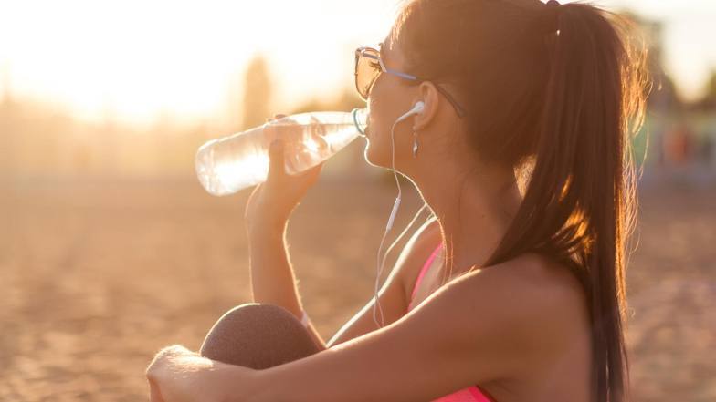 امرأة شابة تان تجلس على الشاطئ في غروب الشمس تشرب زجاجة ماء