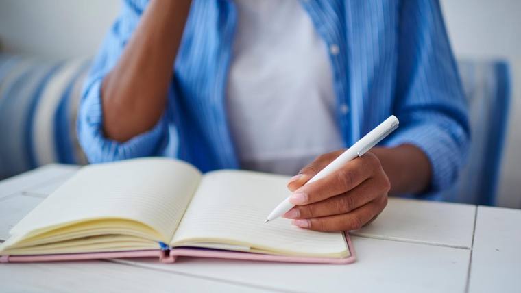صورة مقربة لامرأة ترتدي قميصًا أزرق تجلس على مكتب وتحمل قلمًا وتكتب في كتاب