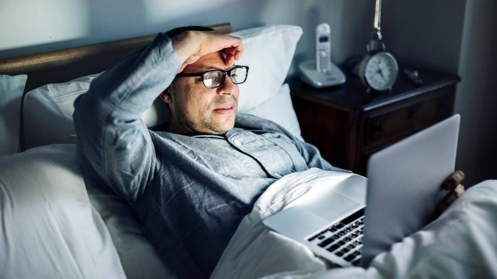 رجل يحك جبهتهُ أثناء استخدامه للكمبيوتر المحمول على السرير. غرفة نومه مظلمة، مضاءة بوهج شاشة الكمبيوتر المحمول.
