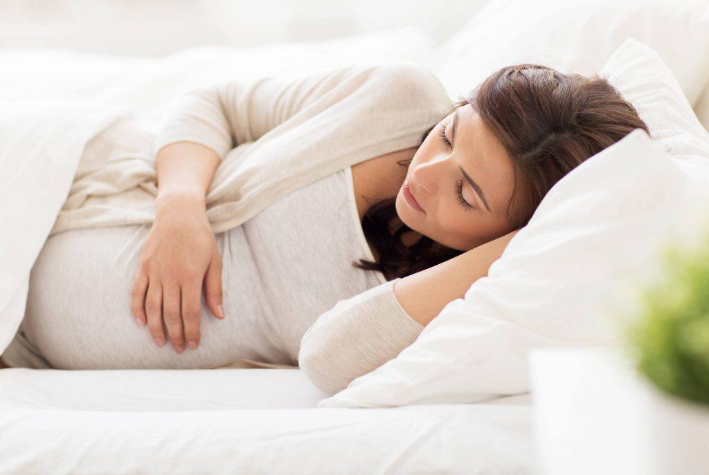 Uma mulher grávida com seus olhos fechados, dormindo na cama com lençóis e travesseiros brancos, apoiando sua mão no seu estômago