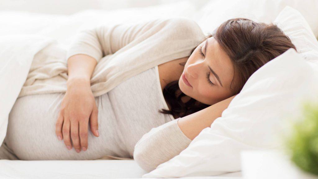 Uma mulher grávida com seus olhos fechados, dormindo na cama com lençóis e travesseiros brancos, apoiando sua mão no seu estômago