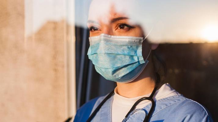 profissional da área médica do sexo feminino, talvez uma enfermeira, usando uma máscara e parecendo triste, séria, preocupada.