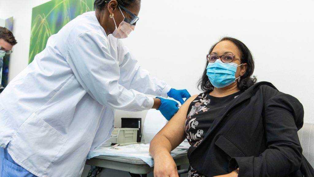 uma enfermeira negra em EPI administrando uma vacina COVID-19 a um funcionário, talvez uma mulher latina em uma jaqueta preta sendo vacinada