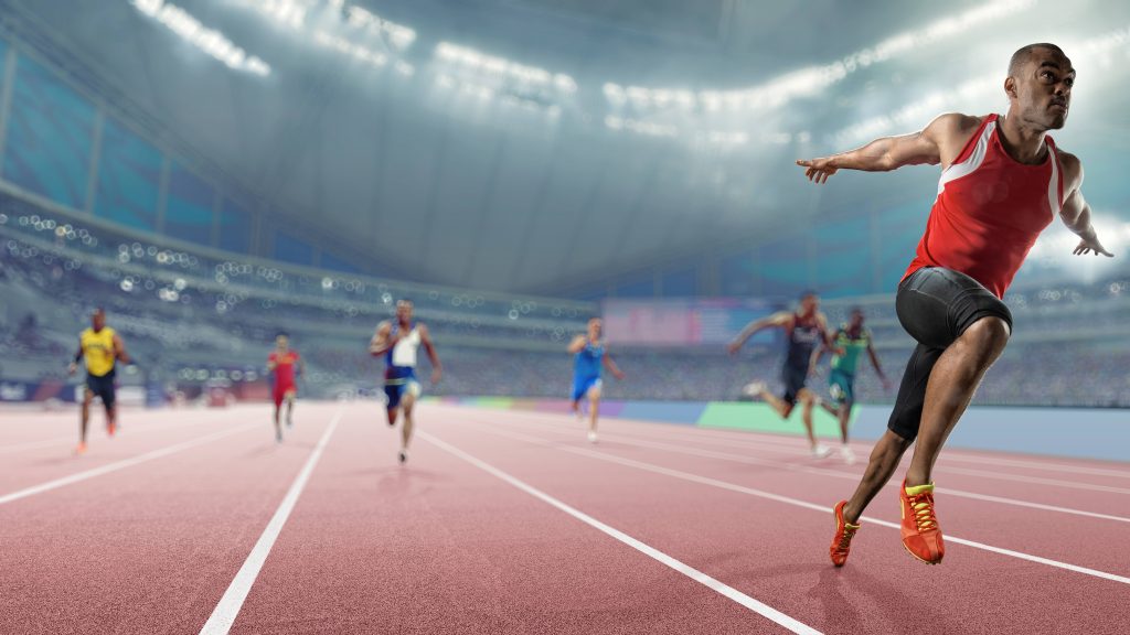 Imagem de grande angular e profundidade de campo de um atleta profissional de pista masculina correndo sobre a linha de chegada de uma corrida com os braços estendidos em vitória com expressão composta. O atleta está disputando uma prova de sprint indoor em pista genérica, e termina à frente de seus competidores.