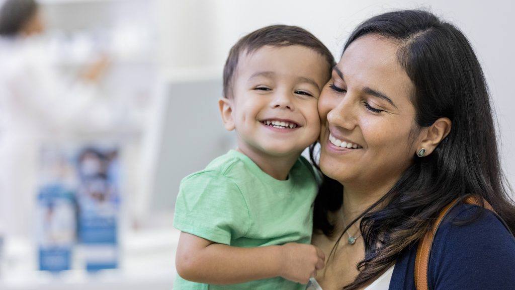 Uma criança sorri enquanto aperta as bochechas com sua mãe na loja. Sua mãe sorri e fecha os olhos satisfeita.
