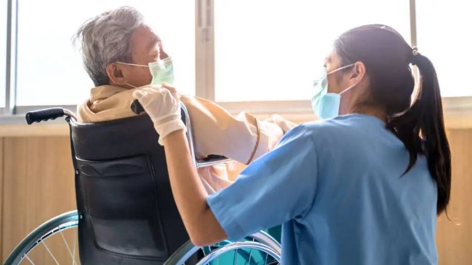 um homem idoso, usando máscara e sentado em uma cadeira de rodas próximo à uma janela com uma profissional de saúde, uma enfermeira usando EPI contra COVID-19