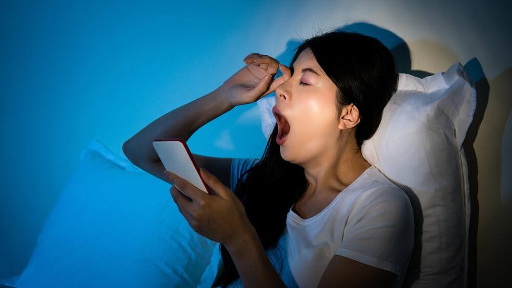 Uma mulher esfrega os olhos enquanto boceja e segura seu smartphone. Ela está encostada em um travesseiro enquanto descansa na cama.