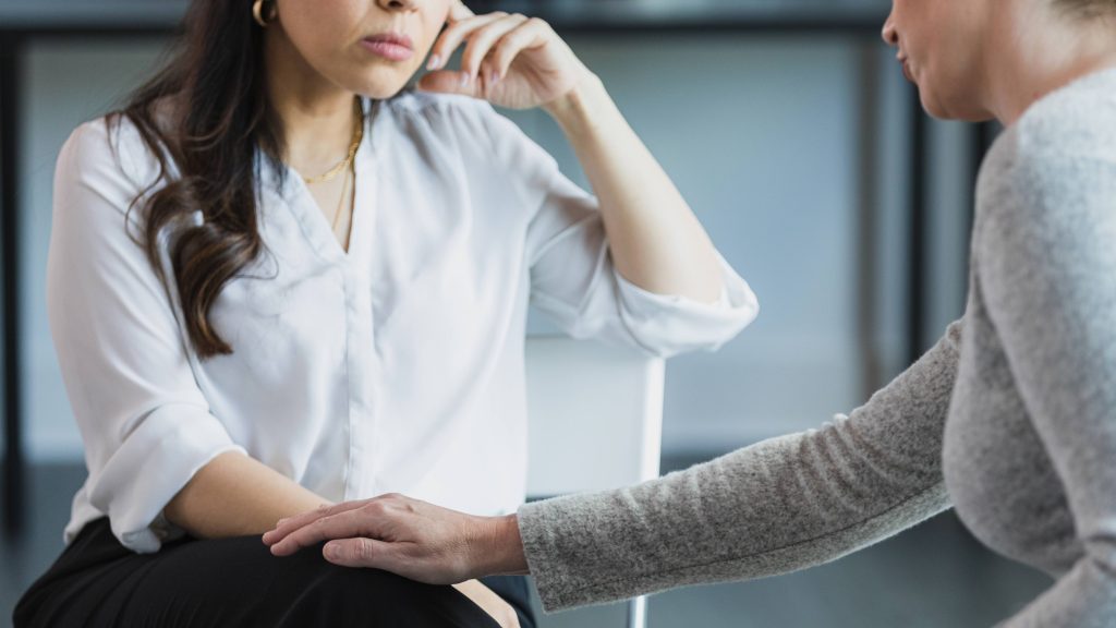 Uma terapeuta de meia idade que não pode ser reconhecida estende a mão para confortar uma mulher de meia idade enquanto elas conversam sobre questões difíceis.
