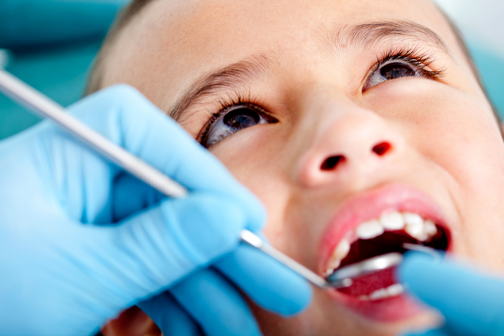Young boy with mouth open and dentist in blue gloves is examining his mouth with dental instruments