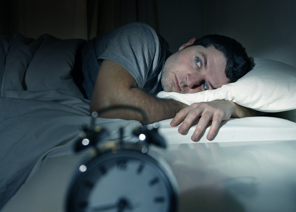 a man in bed in a dark room, with his eyes wide open, looking frustrated, possibly suffering from insomnia or a sleep disorder, with an alarm clock in the foreground