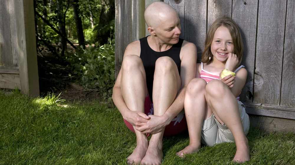 Una madre que lucha contra el cáncer de mama sentada afuera con su hija pequeña junto a una cerca de madera  