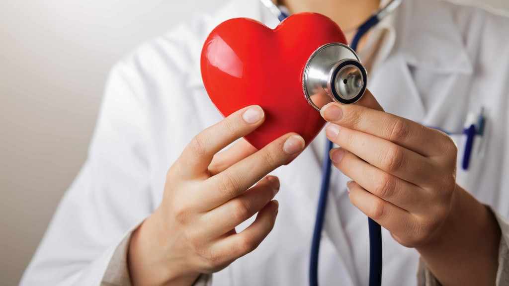 Un proveedor de atención médica con un mandil blanco y un estetoscopio sostiene un corazón de plástico rojo.