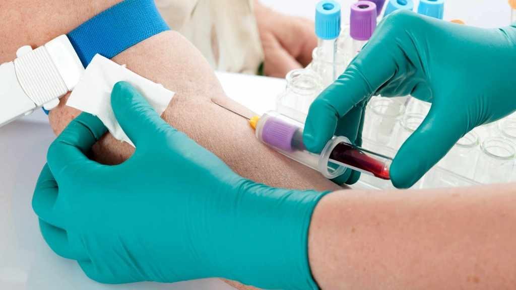 gloved hands of a health care provider using a needle to draw blood from the arm of a patient