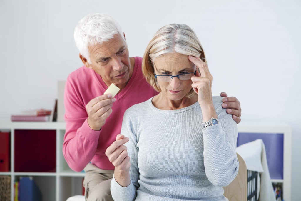 an older man showing a piece of paper to an older woman who is touching her head and trying to remember something