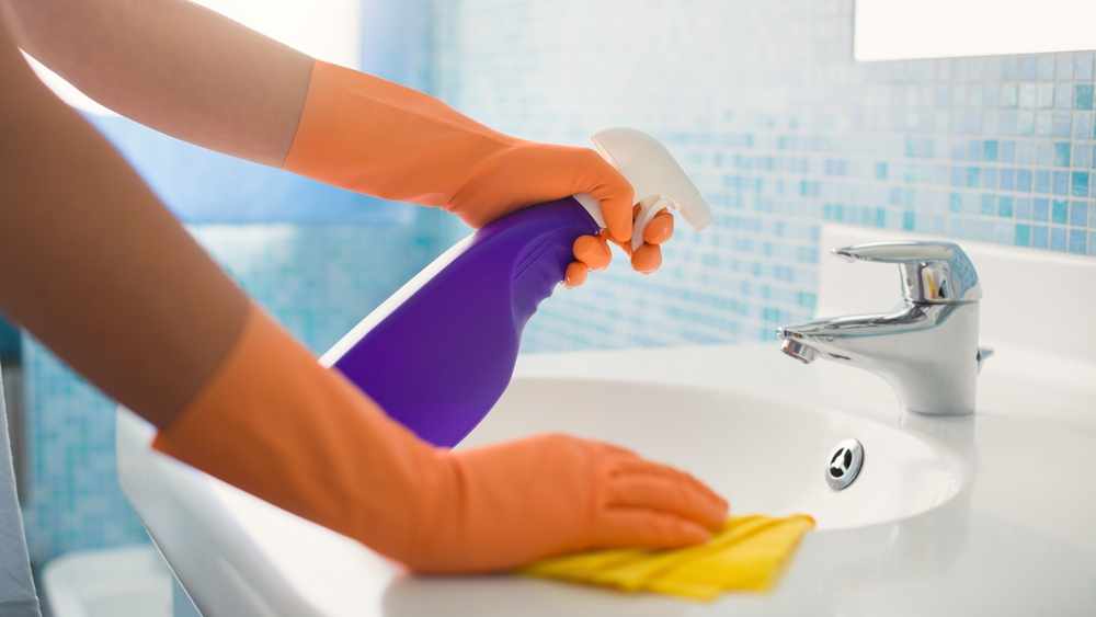 gloved hands spraying cleaner on a bathroom sink faucet and wiping with a cleaning rag