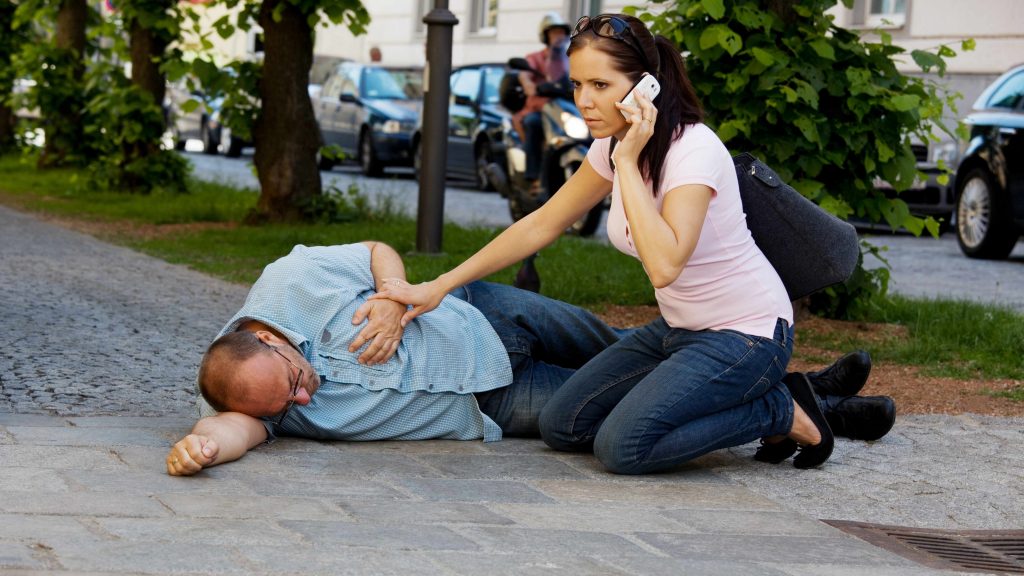 a man lying on the sidewalk holding his chest, having a heart attack or stroke, with a woman helping him and calling for help on the phone