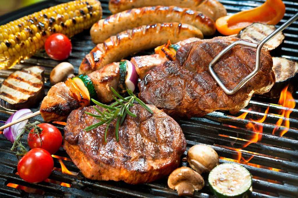 steak, meat, brats and vegetables cooking over a barbecue grill