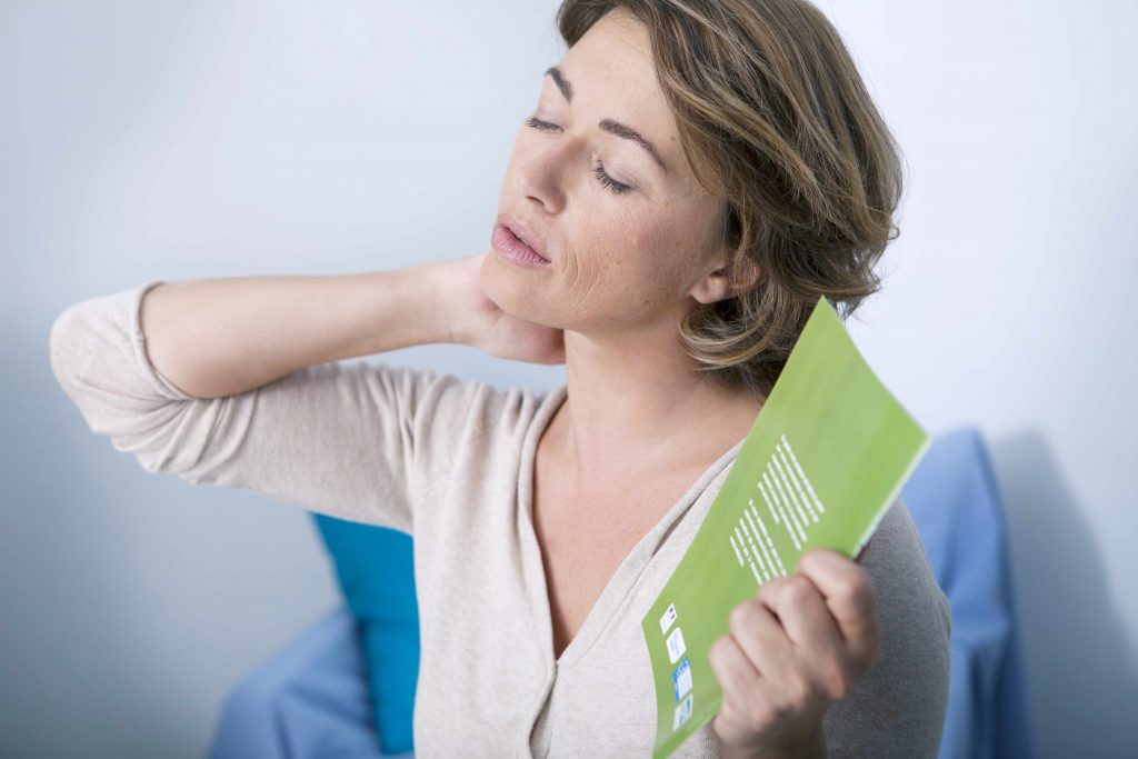 a middle-aged woman rubbing her neck and fanning herself, perhaps sweating with menopausal hot flashes
