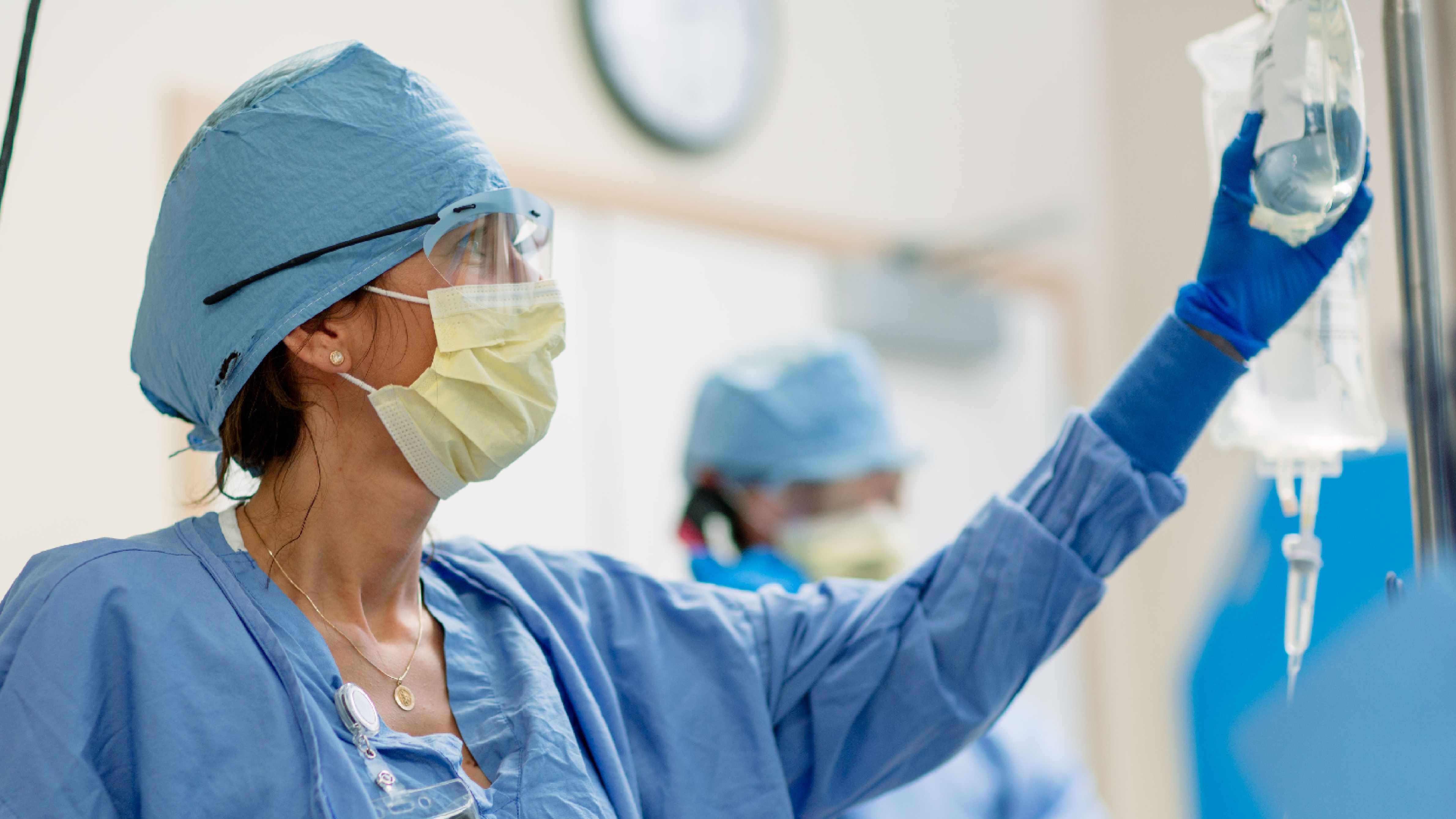 Una enfermera vestida con ropa estéril azul levanta el brazo para alcanzar el suero