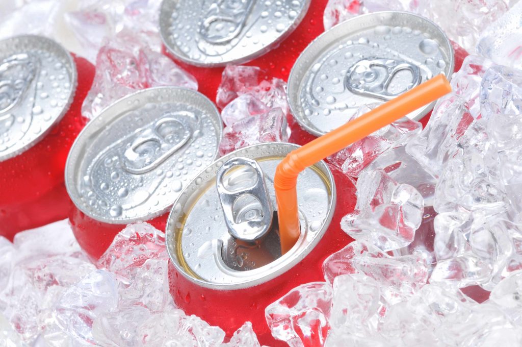 the tops of several cans of soda, packed in ice, one opened and with an orange straw in the top