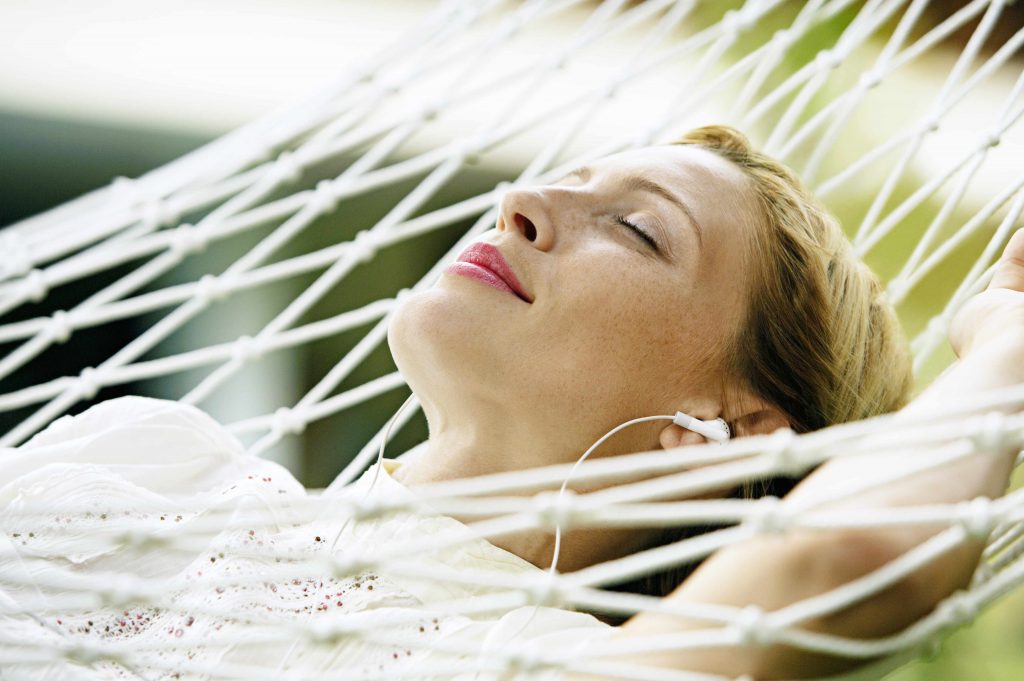 a close-up of a woman relaxing in a hammock, eyes closed, smiling, with earbuds in her ears