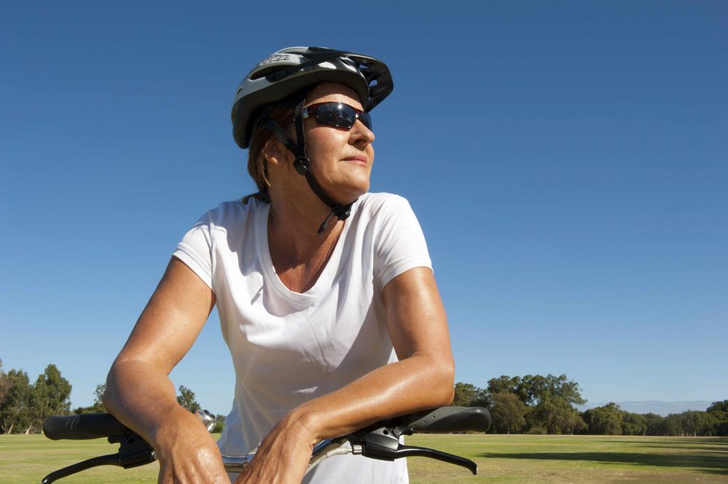 a woman outdoors on a sunny day, with a bike, wearing a helmet and sunglasses, sweating