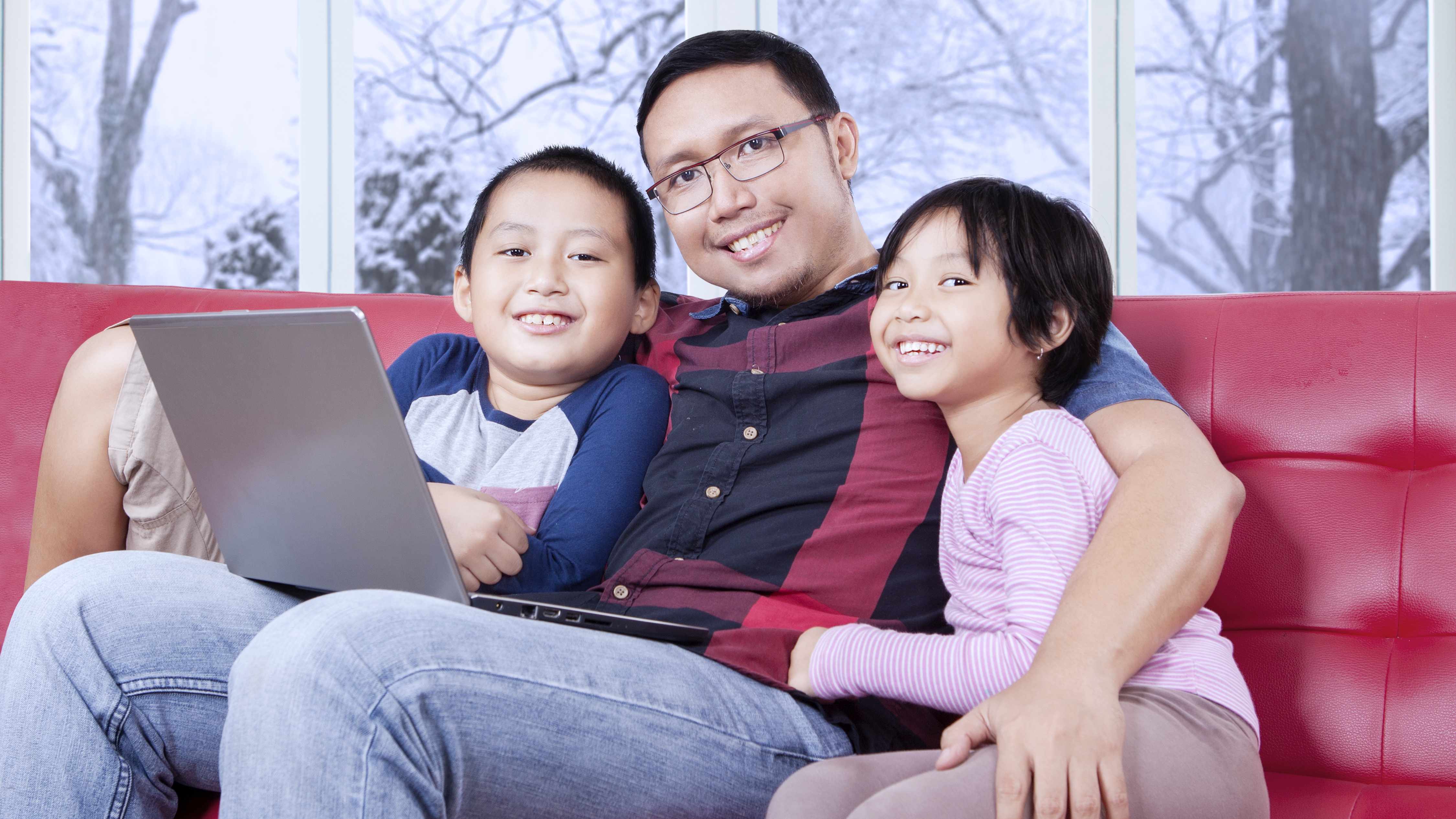 Un joven padre sentado en un sofá con sus hijos pequeños y una computadora portátil sobre las piernas sonríe y luce relajado