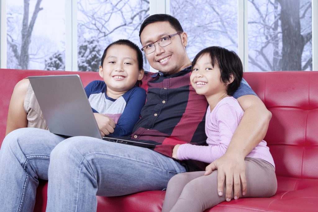 a young father, with a young son and daughter, sitting on a couch, with a laptop on his lap, smiling and relaxed
