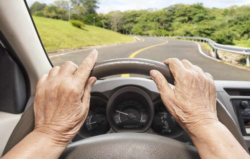 a close-up of an older woman's hands on a steering wheel and the view of the road ahead through the car windshield