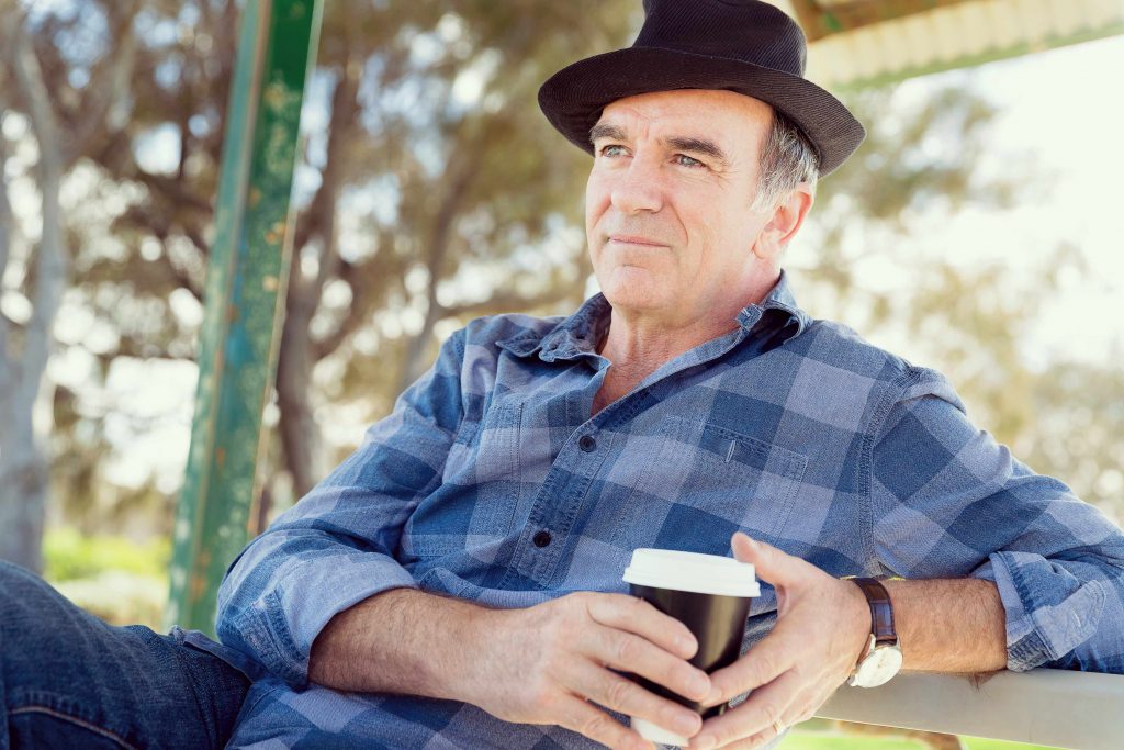 an older man sitting outside with a cup of coffee in his hands and a hat on his head