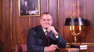 Male sitting at a dark wood desk looking toward the viewer dressed in a pinstriped suit, with his hand near his chin