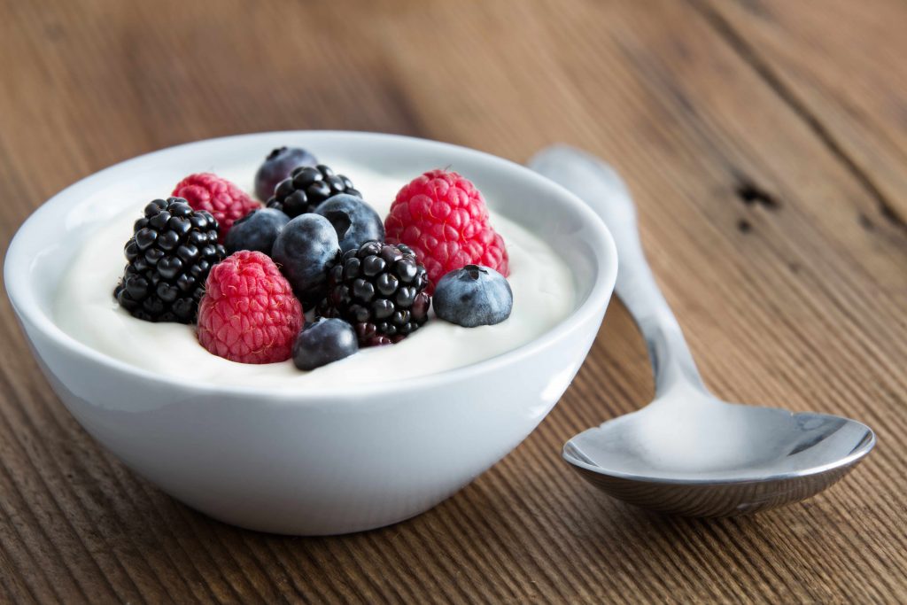 a bowl of yogurt topped with fresh berries, set on a wooden table alongside a large silver spoon