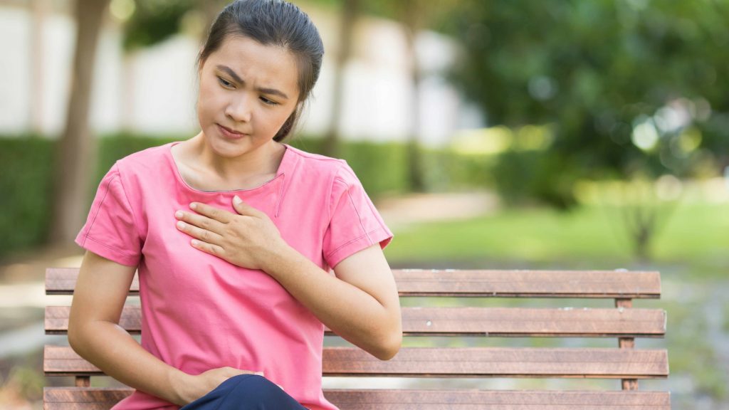 Una mujer joven y sentada en la banca de un parque se sostiene el pecho porque siente acidez estomacal o molestias