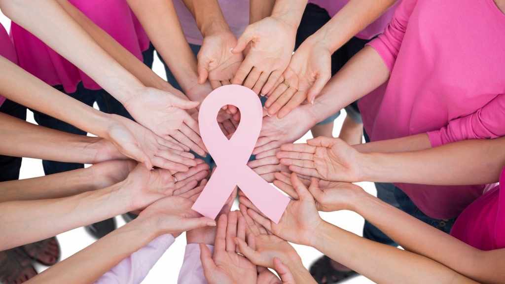 a circle of women in pink shirts, with hands gathered in the middle holding a pink ribbon symbolizing breast cancer awareness