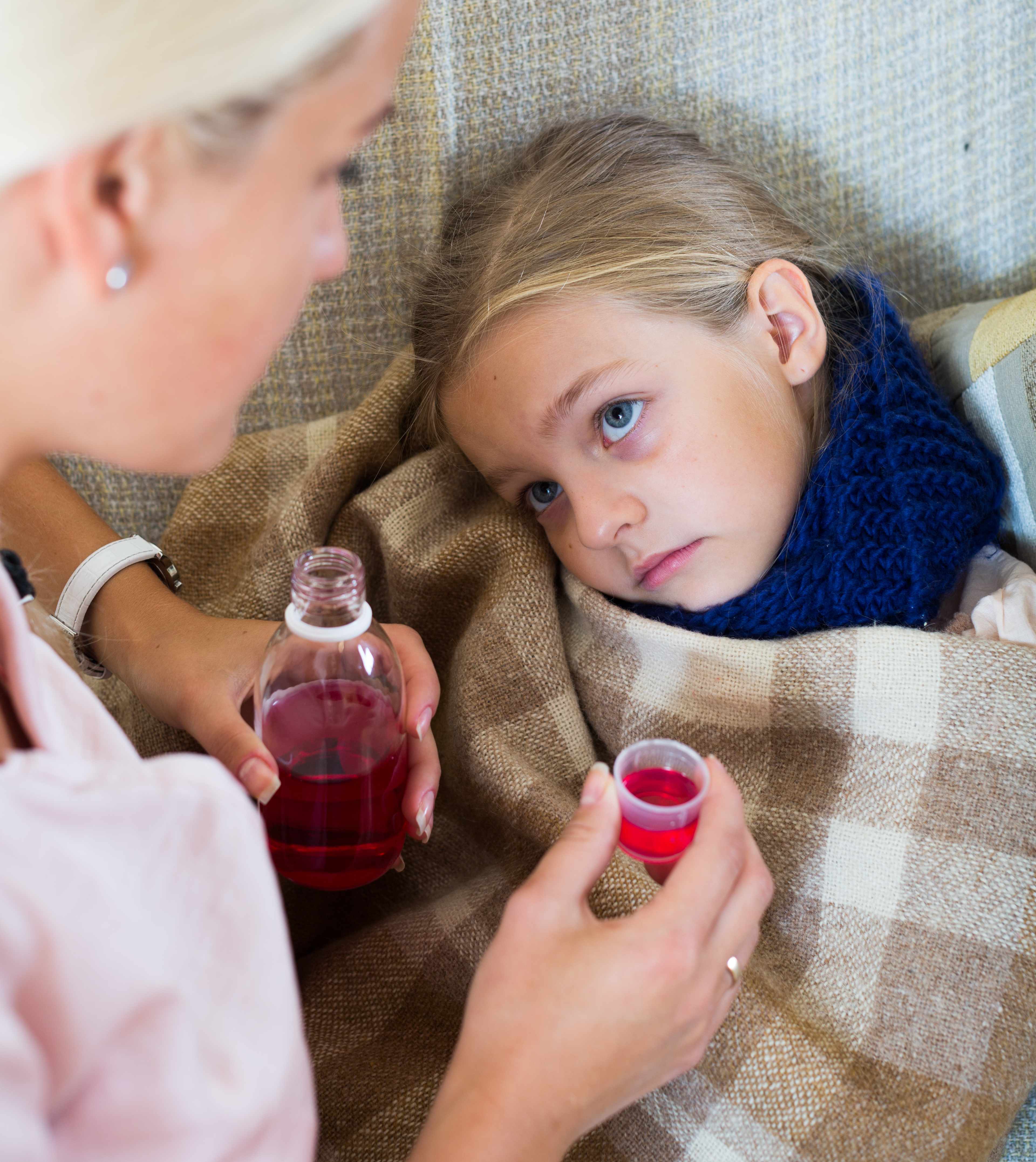 a little girl looking sick and sad with adult trying to give her cough medicine