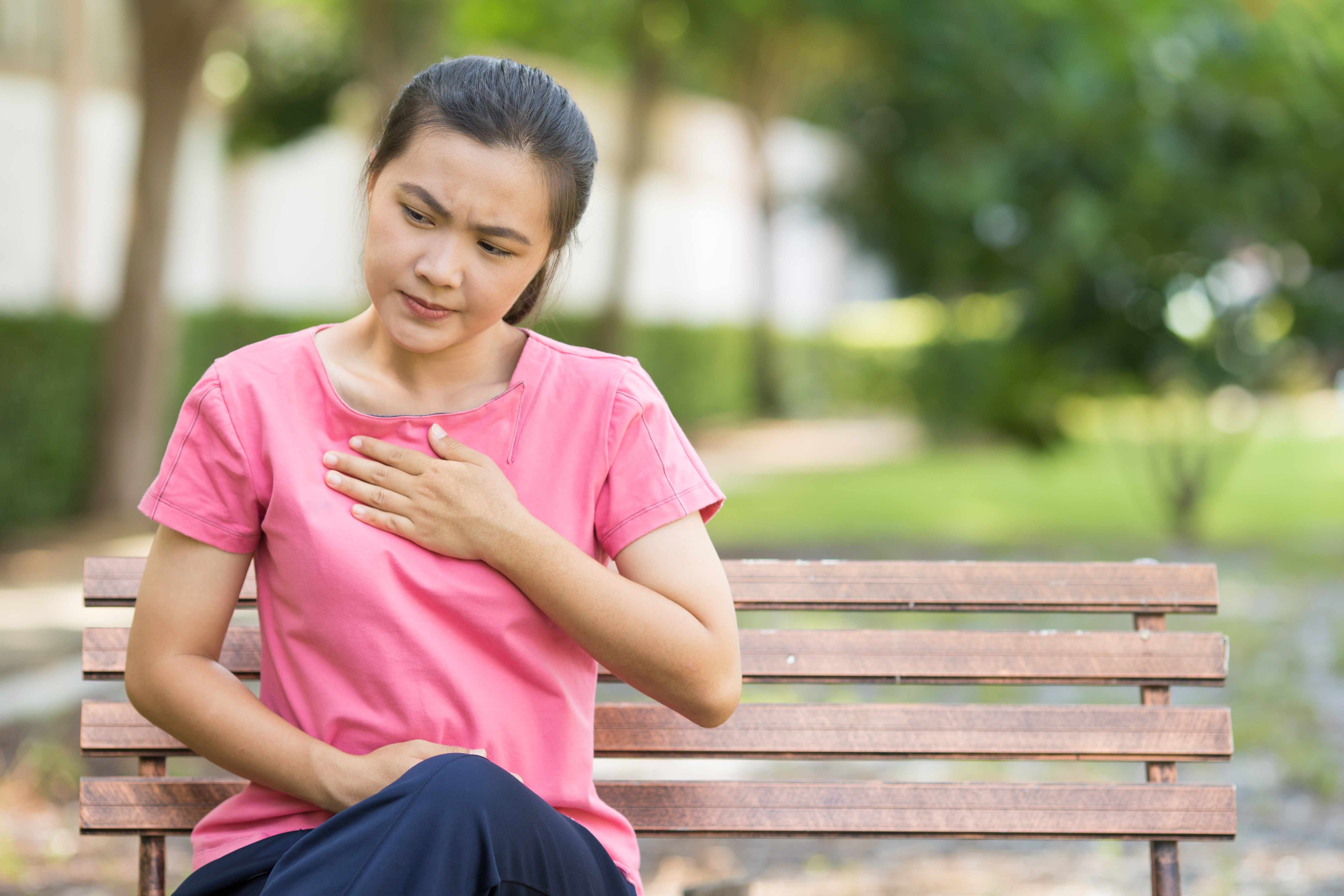 a young woman sitting on a park bench holding her chest because of heartburn or discomfort
