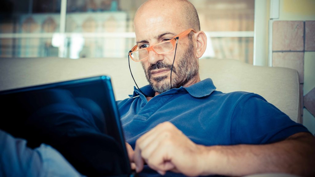Hombre de mediana edad, calvo, con lentes y sentado en un sofá mientras mira a la pantalla de una computadora portátil