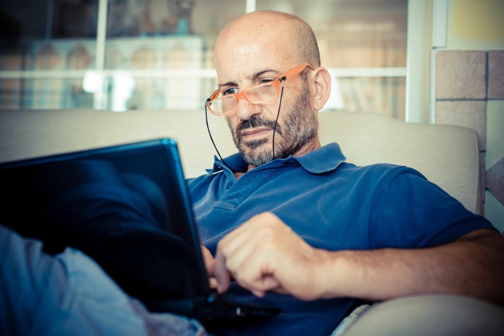 a bald middle aged man with glasses, sitting on a couch, looking at a laptop screen