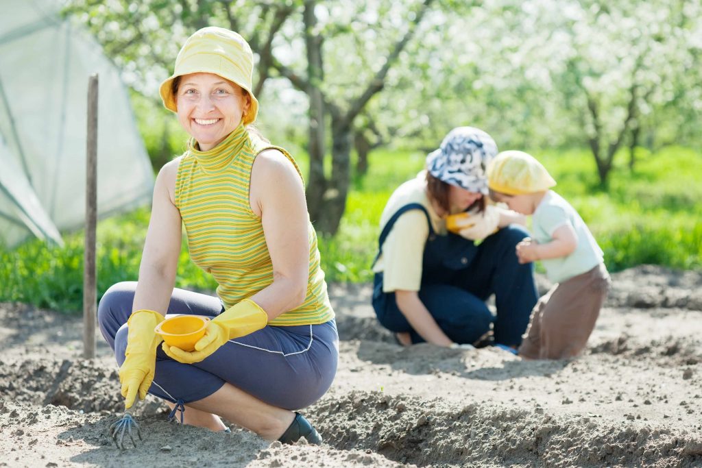 a smiling woman wearing a hat, crouched down, working in a garden, with a younger woman and child in the background