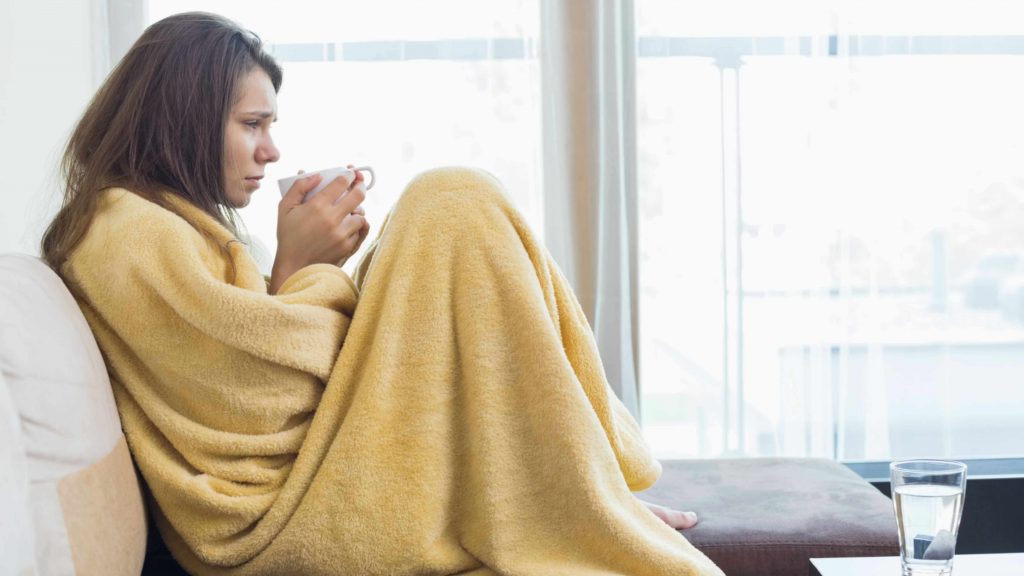 a woman wrapped in a blanket looking sick with cold or flu, holding a cup