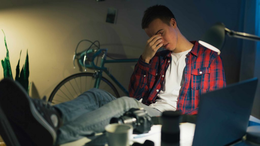 a young teen or college student sitting at his computer rubbing his eyes tired and exhausted
