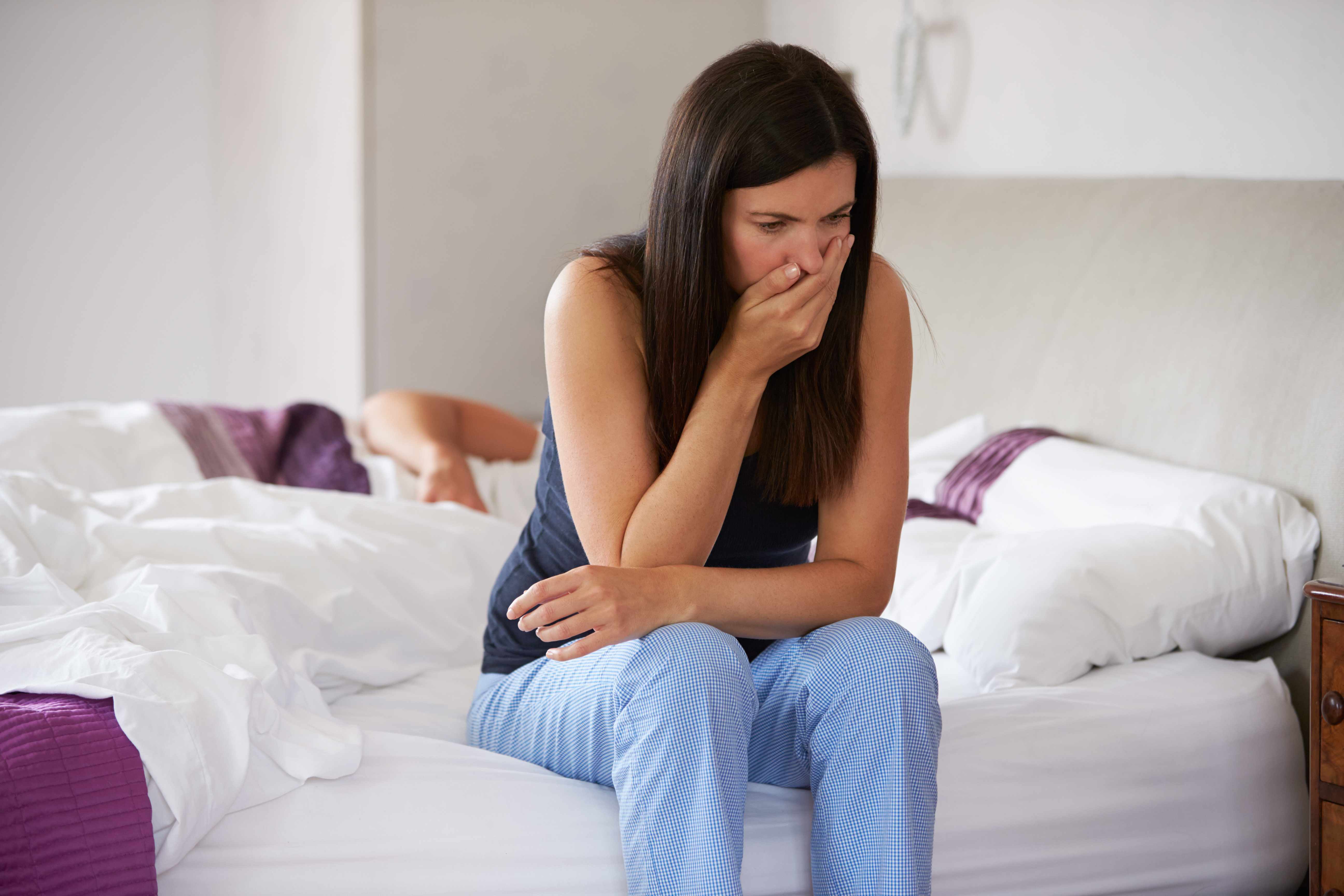 a woman sitting on the edge of a bed with her hand over her mouth looking like she has morning sickness