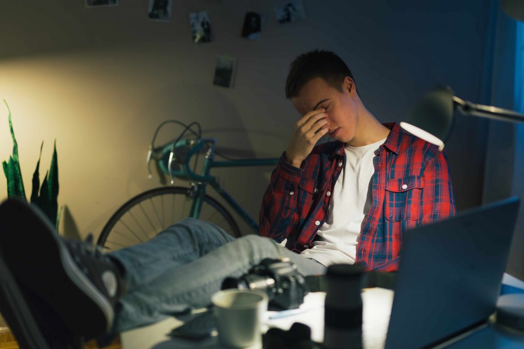 a young teen or college student sitting at his computer rubbing his eyes tired and exhausted