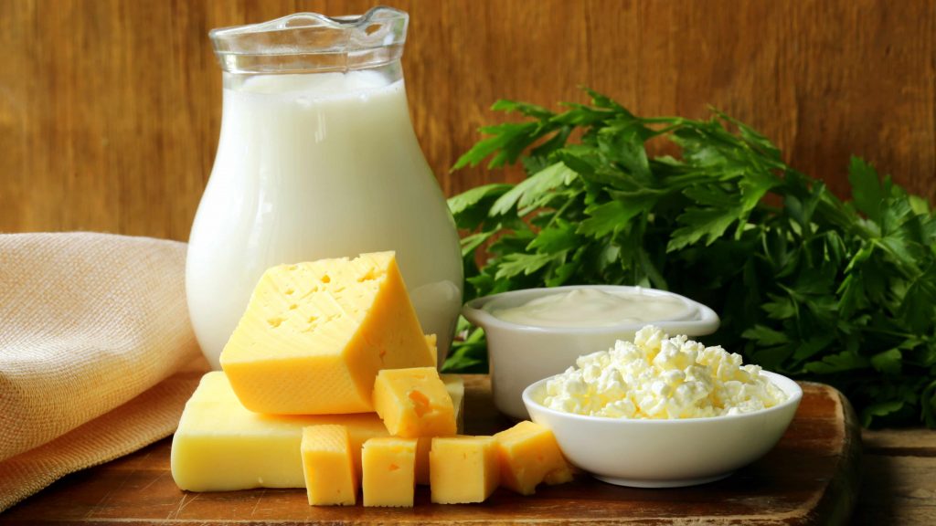 Imagen de productos lácteos, como leche, queso, crema agria y requesón, arreglados sobre una tabla de cortar y decorados con hojas verdes al fondo