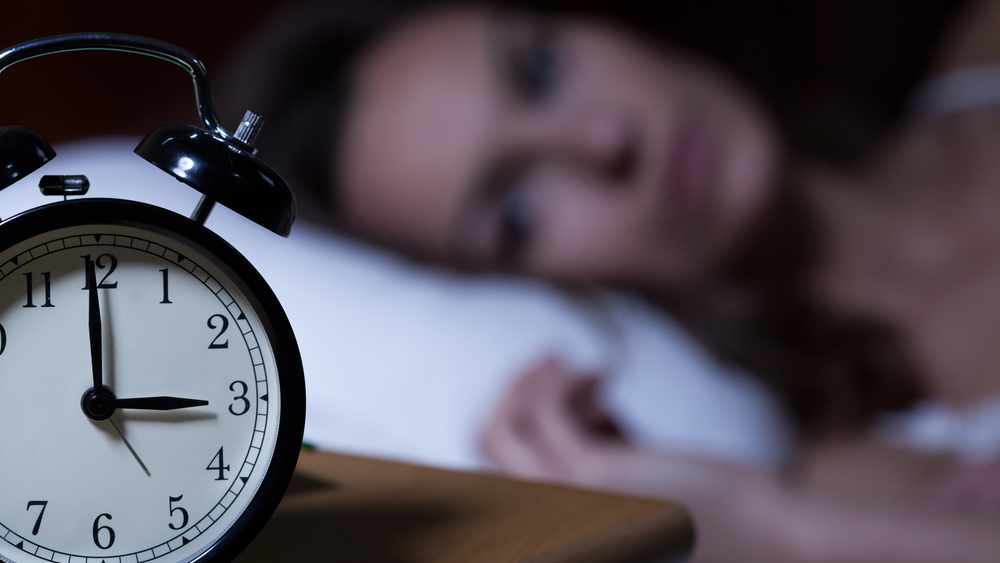 a close-up of an alarm clock reading 3 o'clock, with a wide awake woman lying on a pillow in the background, out of focus, suffering from insomnia