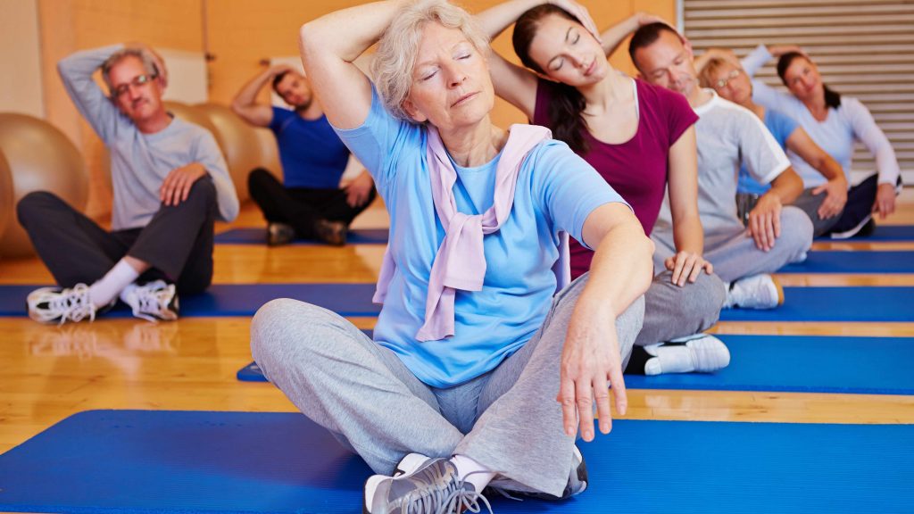 Un grupo de personas, jóvenes y viejas, hombres y mujeres, sentados sobre esteras de yoga en un gimnasio y estirándose