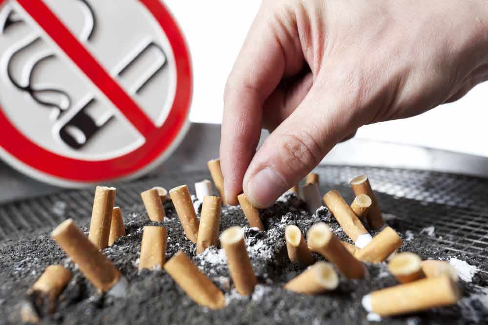 an ashtray full of cigarette butts in front of a no-smoking sign, with a hand shoving another butt into the pile