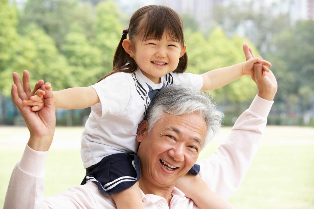 a close-up of a smiling grandfather and granddaughter, outdoors on a sunny day, with her riding on his shoulders