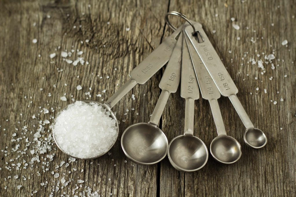 a set of silver measuring spoons on a rough wooden surface, the largest filled with salt spilling over the edges